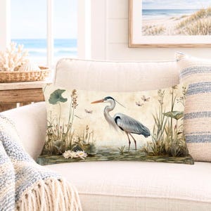 May include: A rectangular throw pillow featuring a detailed illustration of a grey heron standing in a marshy landscape with water lilies, tall grasses, and small flying insects against a soft, muted beige background with a vintage aesthetic.