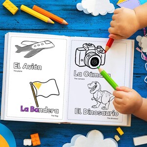 May include: A child colours in a Spanish language colouring book with pictures of a plane, camera, flag, and dinosaur. The book has the words "El Avión", "La Cámara", "La Bandera", and "El Dinosaurio" written on it.