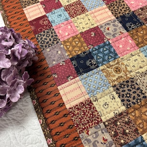 May include: A patchwork quilt featuring a grid of small squares in various patterns and colors including blue, red, brown, and cream. The quilt is bordered by a wide strip of orange fabric with a dark brown geometric pattern and a floral edge.