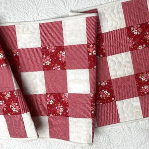 Może przedstawiać: Czerwono-biały bieżnik na st&oacute;ł w kratę z pikowanym wzorem. Bieżnik ma naprzemienne kwadraty z czerwonej i białej tkaniny, z kt&oacute;rych niekt&oacute;re mają wz&oacute;r kwiatowy. Bieżnik jest wykonany z tkaniny.