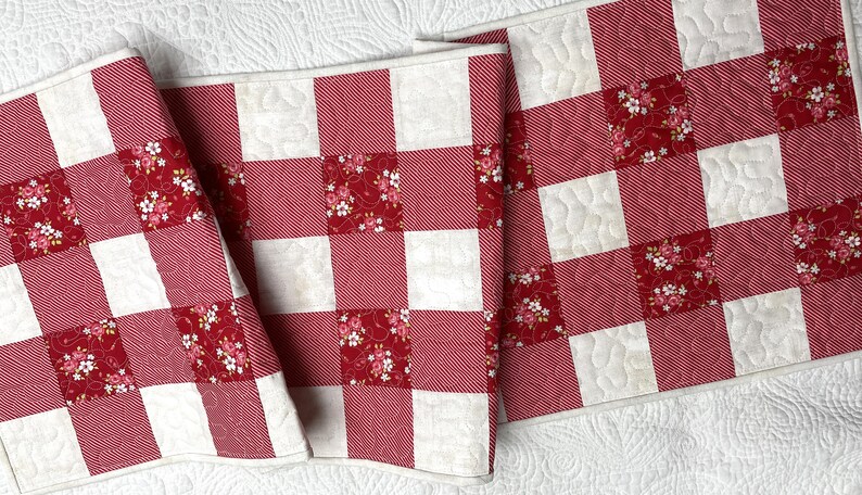 Może przedstawiać: Czerwono-biały bieżnik na st&oacute;ł w kratę z pikowanym wzorem. Bieżnik ma naprzemienne kwadraty z czerwonej i białej tkaniny, z kt&oacute;rych niekt&oacute;re mają wz&oacute;r kwiatowy. Bieżnik jest wykonany z tkaniny.