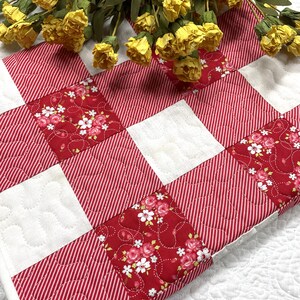 Może przedstawiać: Pikowany bieżnik na st&oacute;ł z czerwono-białym wzorem w paski i kwiatowymi akcentami. Bieżnik ma wz&oacute;r patchworkowy z naprzemiennymi kwadratami czerwonych pask&oacute;w i kwiatowej tkaniny. Ż&oacute;łte kwiaty ułożone na wierzchu.