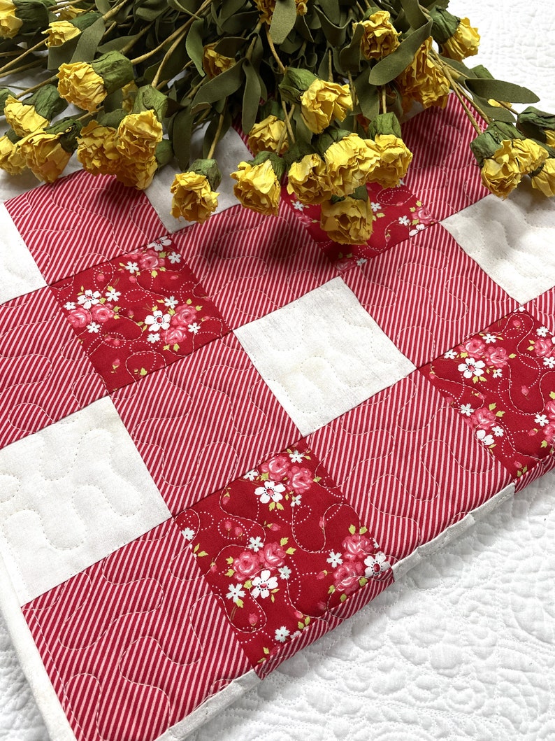 Może przedstawiać: Pikowany bieżnik na st&oacute;ł z czerwono-białym wzorem w paski i kwiatowymi akcentami. Bieżnik ma wz&oacute;r patchworkowy z naprzemiennymi kwadratami czerwonych pask&oacute;w i kwiatowej tkaniny. Ż&oacute;łte kwiaty ułożone na wierzchu.