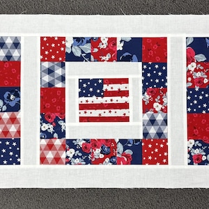 May include: A patriotic table runner featuring three American flags in the center, surrounded by red, white, and blue patchwork squares. The runner is bordered with a white fabric, creating a decorative accent for a table.