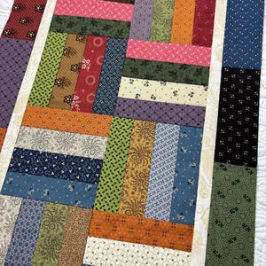 May include: A rectangular table runner featuring a patchwork design composed of numerous small, vertical and horizontal fabric strips. The quilt-style pattern displays a variety of floral, geometric, and dotted prints in shades of red, blue, green, and gold.