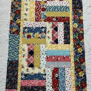 May include: A rectangular table runner featuring a patchwork design of various fabric strips in red, blue, yellow, and white. The pattern includes floral, polka dot, plaid, and striped motifs, framed by a yellow border and a floral outer edge.