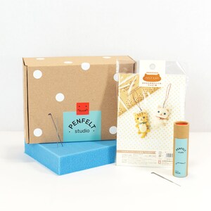 Pu&ograve; includere: Un kit per la creazione di ornamenti di gatto in feltro. Il kit include una scatola marrone con pois bianchi, un sacchetto di plastica trasparente con istruzioni e pezzi di feltro, un tubo di cartone marrone con aghi per infeltrire e un cuscino di schiuma blu. Le istruzioni includono il testo "HAMANAKA FELT CAT" and "あみぐるみをつくって、プレゼントに。"