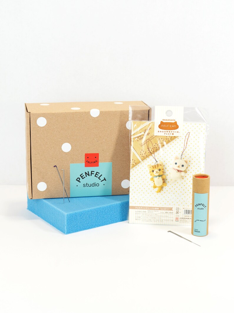 Pu&ograve; includere: Un kit per la creazione di ornamenti di gatto in feltro. Il kit include una scatola marrone con pois bianchi, un sacchetto di plastica trasparente con istruzioni e pezzi di feltro, un tubo di cartone marrone con aghi per infeltrire e un cuscino di schiuma blu. Le istruzioni includono il testo "HAMANAKA FELT CAT" and "あみぐるみをつくって、プレゼントに。"