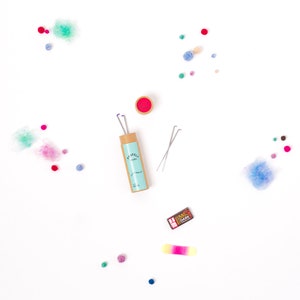 Pu&ograve; includere: Un set di aghi per infeltrire in un tubo di cartone con un'etichetta blu e bianca che dice "PUMPKIN" e "Studio". Gli aghi sono accanto a un piccolo contenitore rosso e una piccola scatola marrone e dorata che dice "85% DARK". Ci sono anche piccole palline di feltro colorato sparse intorno agli aghi.