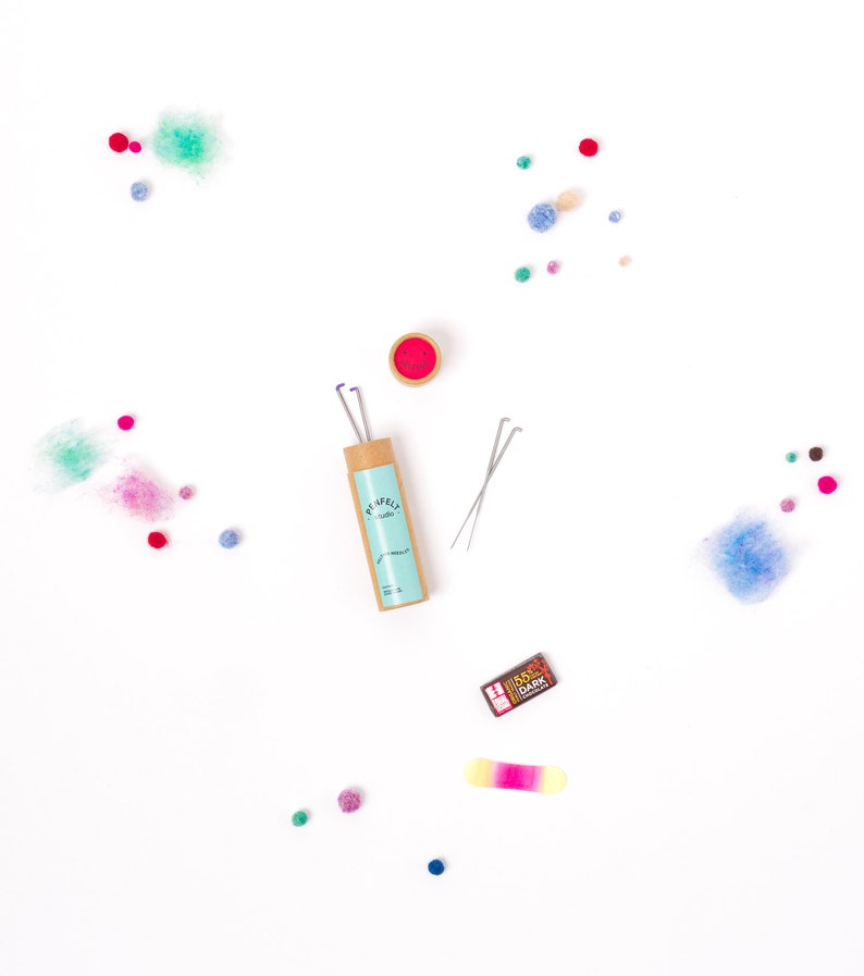 Pu&ograve; includere: Un set di aghi per infeltrire in un tubo di cartone con un'etichetta blu e bianca che dice "PUMPKIN" e "Studio". Gli aghi sono accanto a un piccolo contenitore rosso e una piccola scatola marrone e dorata che dice "85% DARK". Ci sono anche piccole palline di feltro colorato sparse intorno agli aghi.