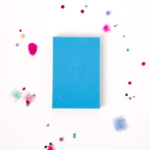 Pu&ograve; includere: Una spugna rettangolare blu su uno sfondo bianco circondata da piccole palline di feltro colorate.