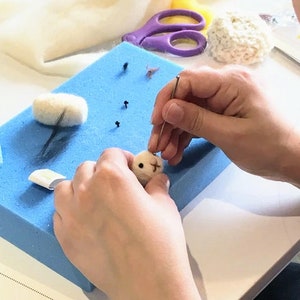 Pu&ograve; includere: Una persona sta usando un ago per infeltrire una piccola testa di animale di lana bianca. La testa ha occhi neri e un piccolo naso rosa. La persona sta lavorando su un tappetino di schiuma blu.