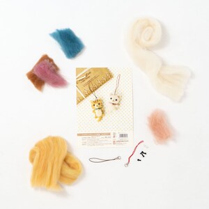 Pu&ograve; includere: Un kit di feltro con lana di vari colori, tra cui giallo, rosa, blu e bianco. Il kit include anche uno spago rosso, due perline nere e un piccolo cartoncino con le istruzioni. Il cartoncino ha il testo "Happy You" e una piccola illustrazione di due gatti.