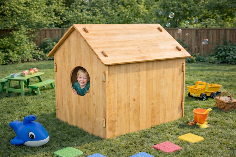 May include: A wooden playhouse with a round window, a child peeking out, and a pitched roof. The playhouse is surrounded by toys, including a yellow dump truck, a blue whale-shaped sprinkler, and a green picnic table. Bubbles float in the air.