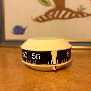 May include: A vintage, cream-colored kitchen timer with a black dial displaying numbers from 5 to 55. The timer has a conical shape and a white indicator. The timer is sitting on a wooden surface.