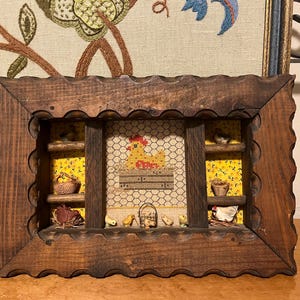 May include: A rustic wooden shadow box with a scalloped frame. Inside, a chicken-themed display features a cross-stitched hen, miniature baskets, and small chicks. The background is yellow fabric with a floral pattern.