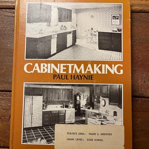 May include: An orange book titled "Cabinetmaking" by Paul Haynie. The cover features black and white images of kitchens. A sticker indicates the subject area is Trade & Industry, and the grade level is High School.