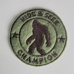 May include: A round, olive green patch with the words "HIDE & SEEK CHAMPION" embroidered in black. The patch features a brown silhouette of Bigfoot in the center, with two black star accents.