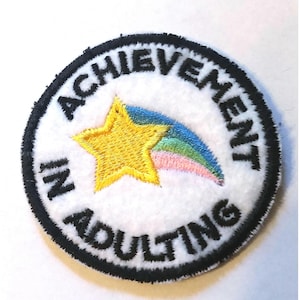 May include: Embroidered patch with the words "ACHIEVEMENT IN ADULTING" in black around a white circle. A yellow star with a rainbow comet tail is in the center. The patch has a black border.