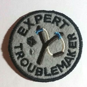 May include: A round, gray patch with black trim and the words "EXPERT TROUBLEMAKER" embroidered in black. A black and white slingshot with blue accents is in the center of the patch. The patch is made of felt.