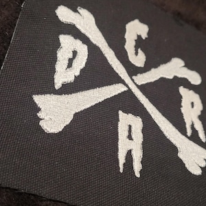 May include: A black fabric patch featuring white embroidered text and bone-shaped graphics. The design consists of two crossed bones forming an X shape, with the letters D, C, A, and R positioned in the four quadrants created by the crossed bones.