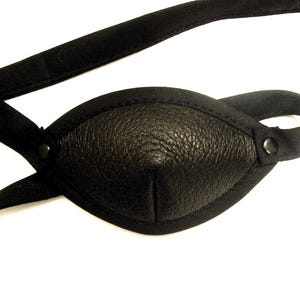 May include: Black leather eye patch with a textured surface and a black fabric strap. The eye patch is oval-shaped and has a stitched border. The strap is adjustable and secured with a button.