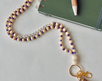 May include: A beaded lanyard with the name "MISS PHEEBE" spelled out in white letter beads. The lanyard features light wood-colored beads and purple accent beads. It includes a gold-colored clasp and a brown tassel.
