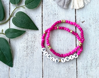 May include: Two pink and gold beaded bracelets with white letter beads spelling out "Girls Trip".