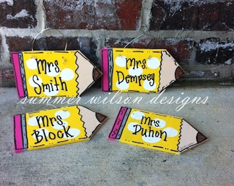 May include: Four yellow pencil-shaped wooden signs with pink and gray accents. Each sign has a white circle with the text "Mrs." and a teacher's last name: Smith, Dempsey, Block, and Duhon.