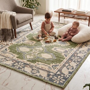 May include: A rectangular area rug featuring a green central field with a cream-colored floral medallion pattern, surrounded by a dark blue border decorated with stylized floral motifs and animal figures in cream and light blue tones.