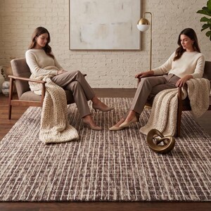 May include: A rectangular area rug featuring a dark brown and cream grid pattern with a textured, woven appearance. The rug is positioned on a dark wood floor, serving as a foundation for two wooden armchairs, each draped with a chunky knit throw.