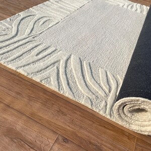 May include: A cream-colored rectangular rug with a textured surface and a rolled-up section. The rug features a raised, abstract design along the border. The rolled section reveals a black backing.