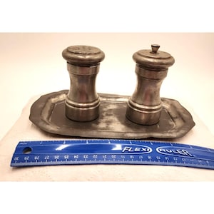 May include: A vintage pewter salt and pepper shaker set with a matching tray. The shakers have a classic, rounded design with a perforated top for the salt and a small knob on the pepper shaker. The tray is rectangular with a slightly scalloped edge.