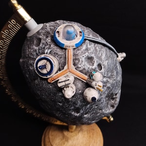 Puede incluir: Un modelo esférico de luna gris y rocoso con una pequeña estación espacial azul y blanca unida a él. La estación espacial tiene una cúpula transparente en la parte superior y una pequeña luz azul en el interior. La luna está sobre una base de madera.