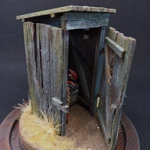 Peut inclure: Modèle réduit d'une cabane en bois avec un aspect rustique et vieilli. La porte est entrouverte, révélant un aperçu de l'intérieur. Le modèle présente des textures détaillées et un petit toit en bois, évoquant une époque révolue.