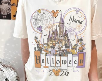 Puede incluir: Una camiseta color crema con un gráfico de un castillo, personajes de dibujos animados y una telaraña dentro de una silueta de orejas de Mickey. El texto dice "Name", "Halloween" y "2026". El diseño incluye tonos naranja, morado y negro.