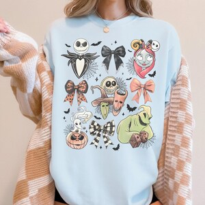 K&ouml;nnte beinhalten: Hellblaues Sweatshirt mit einem Halloween-Motiv. Das Design zeigt Charaktere und Schleifen. Das Design umfasst Jack Skellington, Sally, Oogie Boogie und andere Charaktere aus dem Film. Flederm&auml;use und Spinnweben sind ebenfalls enthalten.