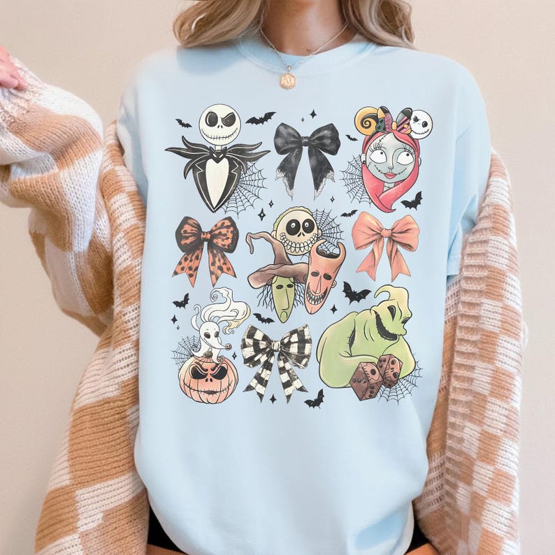 K&ouml;nnte beinhalten: Hellblaues Sweatshirt mit einem Halloween-Motiv. Das Design zeigt Charaktere und Schleifen. Das Design umfasst Jack Skellington, Sally, Oogie Boogie und andere Charaktere aus dem Film. Flederm&auml;use und Spinnweben sind ebenfalls enthalten.