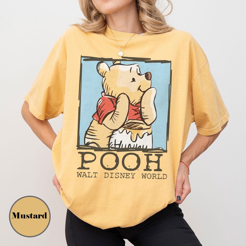 May include: Mustard-coloured t-shirt with a graphic of Winnie the Pooh in a red shirt, with the word "POOH" and "WALT DISNEY WORLD" printed below. The shirt has short sleeves and a relaxed fit.