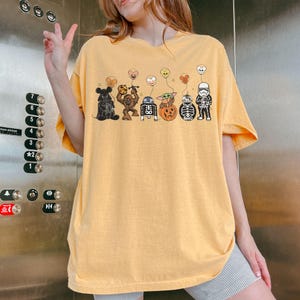 Puede incluir: Camiseta de color amarillo mostaza con una fila de personajes de dibujos animados con globos tem&aacute;ticos de Halloween. Los personajes incluyen un oso, Chewbacca, R2-D2, Baby Yoda, una calabaza, un esqueleto y un Stormtrooper. La camiseta es holgada.
