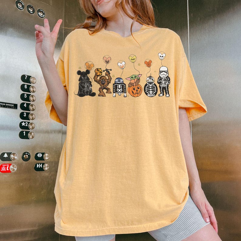 Puede incluir: Camiseta de color amarillo mostaza con una fila de personajes de dibujos animados con globos tem&aacute;ticos de Halloween. Los personajes incluyen un oso, Chewbacca, R2-D2, Baby Yoda, una calabaza, un esqueleto y un Stormtrooper. La camiseta es holgada.