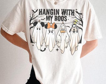 Puede incluir: Camiseta color crema con la frase "HANGIN WITH MY BOOS" y fantasmas de dibujos animados de Mickey Mouse, Minnie Mouse, Pato Donald y Goofy. Los fantasmas cuelgan de un tendedero con murciélagos y estrellas.