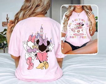 Puede incluir: Camiseta rosa claro con un dibujo de Minnie y Daisy Duck frente a un castillo. La parte delantera de la camiseta tiene un gráfico de Minnie y Daisy en una taza de té con el texto "GIRLS JUST WANNA HAVE FUN!"