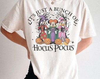 Puede incluir: Camiseta color crema con un diseño temático de Halloween. El diseño incluye personajes de dibujos animados disfrazados de brujas, calabazas y el texto "It's Just a Bunch of Hocus Pocus". La camiseta tiene mangas cortas.
