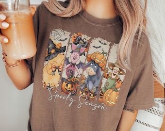 Puede incluir: Una camiseta marrón con un estampado gráfico de cuatro personajes de dibujos animados disfrazados de Halloween, incluyendo una bruja, un murciélago y calabazas. El texto Spooky Season está impreso en blanco bajo las ilustraciones.