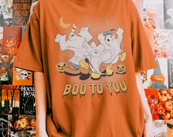 Puede incluir: Una camiseta de color naranja quemado con un gráfico de tres personajes de dibujos animados disfrazados de fantasmas, con zapatos y junto a calabazas. Sobre los personajes hay una luna creciente amarilla y el texto BOO TO YOU.