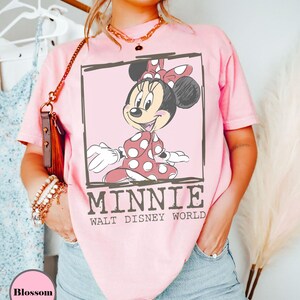 May include: A pale pink t-shirt with a Minnie Mouse graphic in a black outlined box. Minnie is wearing a red polka dot dress and bow. The text "MINNIE WALT DISNEY WORLD" is below the graphic.