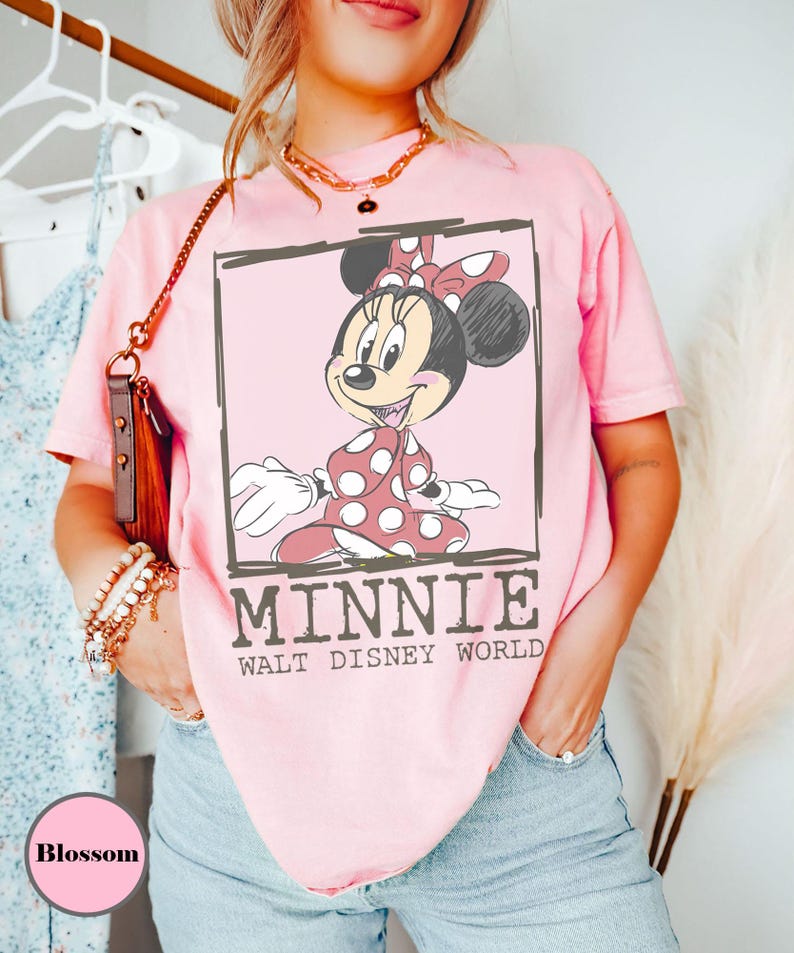 May include: A pale pink t-shirt with a Minnie Mouse graphic in a black outlined box. Minnie is wearing a red polka dot dress and bow. The text "MINNIE WALT DISNEY WORLD" is below the graphic.