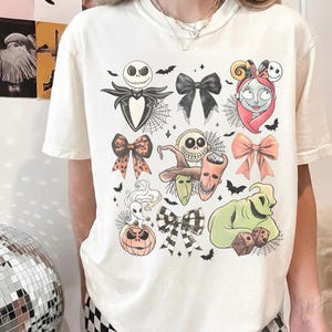 K&ouml;nnte beinhalten: Cremefarbenes T-Shirt mit farbenfrohen Illustrationen von Charakteren und Schleifen aus "The Nightmare Before Christmas". Das Design umfasst Jack Skellington, Sally, Oogie Boogie und andere ikonische Elemente, mit Flederm&auml;usen und Spinnweben.
