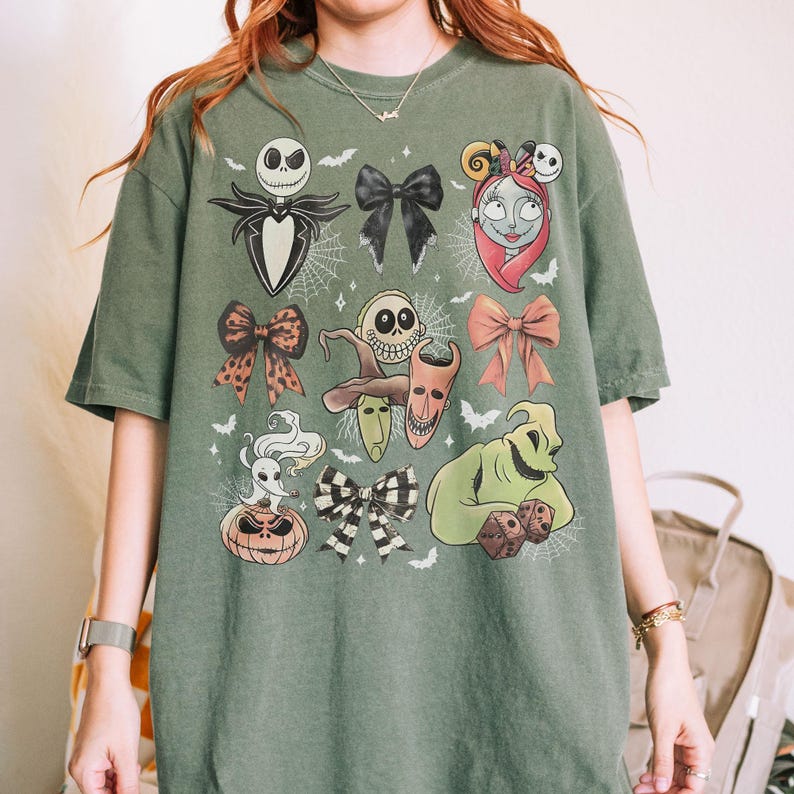 K&ouml;nnte beinhalten: Gr&uuml;nes T-Shirt mit Illustrationen von Charakteren aus The Nightmare Before Christmas. Das Design umfasst Jack Skellington, Sally, Oogie Boogie und andere ikonische Elemente. Das Shirt hat eine lockere Passform und einen Rundhalsausschnitt.